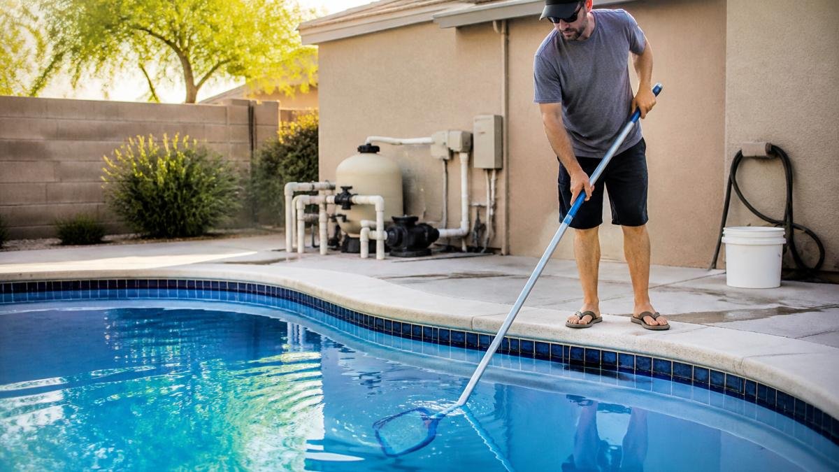 Pool Service Monthly Cost vs DIY: True Price Breakdown 1 Homeowner in casual weekend clothing cleaning a swimming pool with a long-handled brush, with clear blue water and pool equipment visible in the background under morning sunlight